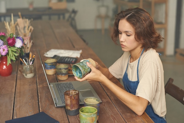 Foto ernste junge kaukasische frau, die mit laptop am holztisch sitzt und keramikbecher in töpfen untersucht...