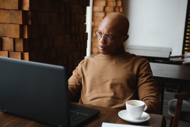 Ernste schwarze männliche Studentenbrille, die Notizen macht, während sie mit Laptop am Tisch sitzt und im gemütlichen Café an einem Projekt arbeitet.