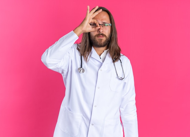 Ernster erwachsener männlicher Arzt, der medizinische Robe und Stethoskop mit Brille trägt