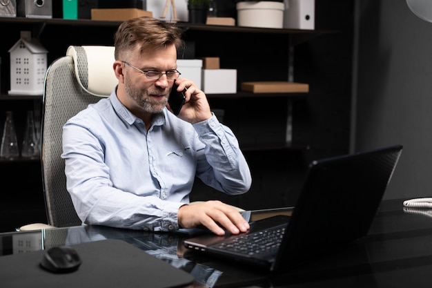 Ernster Geschäftsmann, der an einem Laptop arbeitet und an einem Handy spricht