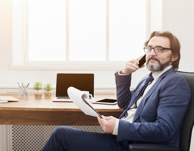 Ernster Geschäftsmann hat Handygespräche und Kaffee am Arbeitsplatz im modernen weißen Bürointerieur, Seitenansicht, Kopierraum