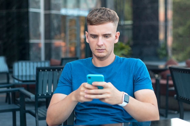 Ernster gutaussehender Kerl, junger konzentrierter trauriger besorgter Mann, der sein Handy des Mobiltelefons betrachtet, Nachricht in der Terrasse im Café draußen tippend. Social Media Sucht. Gadgets, Gerätetechnologiekonzept