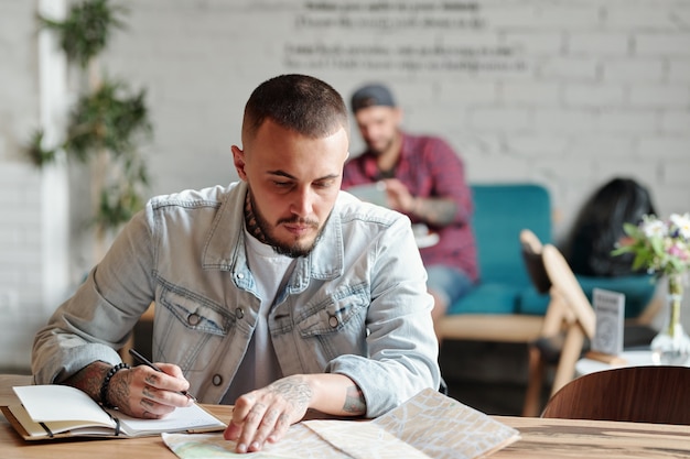 Ernster Hipster-Tourist mit Bart, der am Tisch im Café sitzt und Papierkarte verwendet, während Route plant