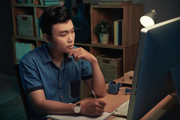 Ernster junger asiatischer Mann, der am Holztisch sitzt und Notizen im Skizzenblock macht, während er spät zu Hause mit dem Computer arbeitet