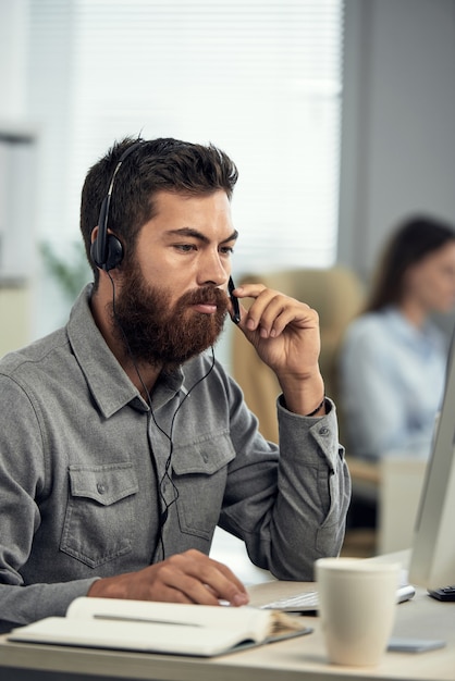 Ernster junger bärtiger Callcenter-Betreiber in Headset mit Mikrofon, der am Schreibtisch sitzt und Laptop verwendet