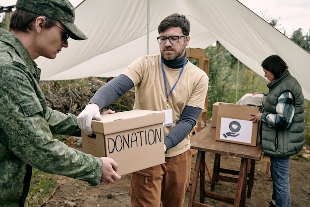 Ernster junger bärtiger Sozialarbeiter mit Abzeichen, der im Freien eine Spendenbox an einen Soldaten weitergibt