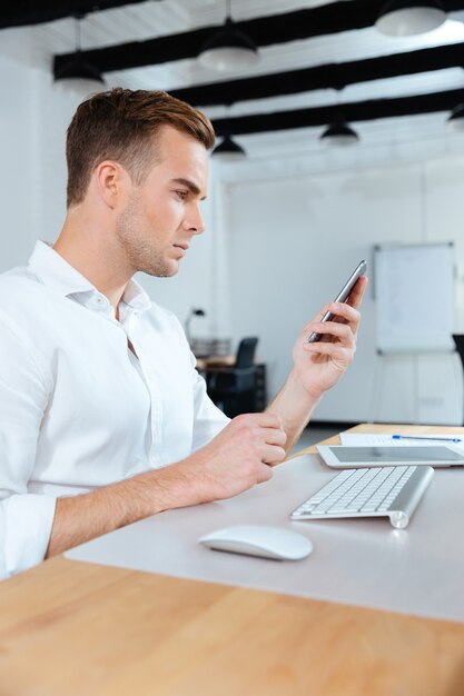 Ernster junger Geschäftsmann, der am Arbeitsplatz sitzt und Handy benutzt