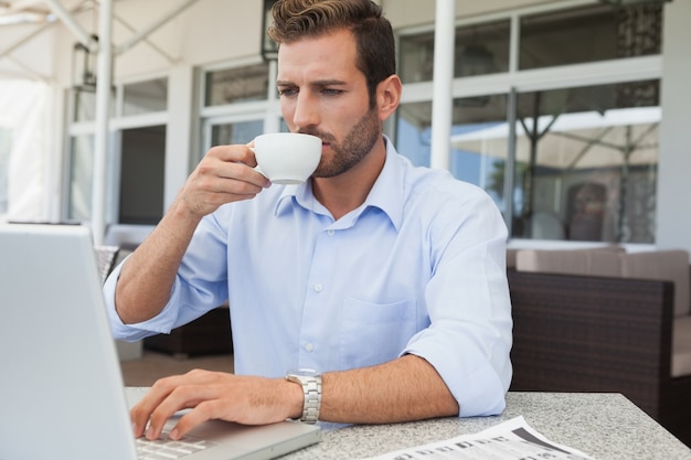 Ernster junger Geschäftsmann, der am trinkenden Kaffee des Laptops arbeitet