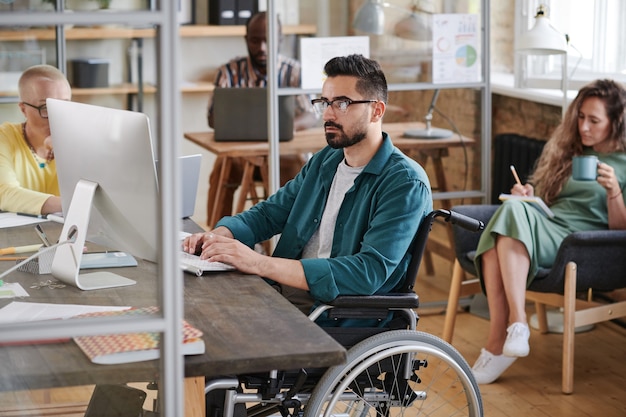 Ernster junger Geschäftsmann im Rollstuhl, der am Tisch sitzt und am Computer tippt, den er mit seinen Kollegen im Büro arbeitet