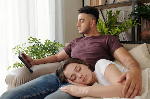 Ernster junger Mann sitzt auf dem Sofa und schaut sich einen Film auf dem Tablet-Computer an, wenn die Freundin schläft ...