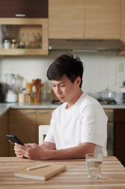 Ernster Mann, der am Küchentisch sitzt und Textnachrichten auf dem Smartphone überprüft