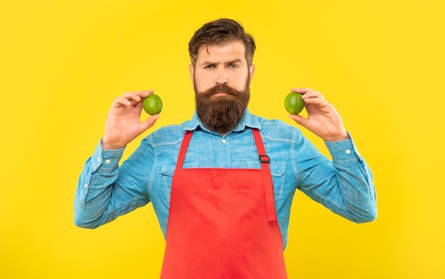 Ernster Mann in roter Schürze mit frischen Limetten Zitrusfrüchten auf gelbem Hintergrund Fruiterer
