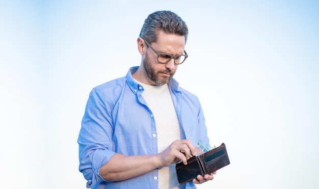 Ernster Mann mit Brille, der Bargeld aus dem Himmelshintergrund der Brieftasche nimmt