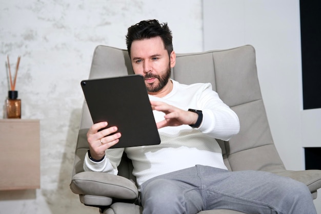 Ernster Mann mit Tablet-Laptop-Computer-Pad, der zu Hause in einem bequemen Sessel sitzt und liest