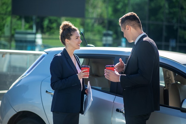 Ernster Mann und lächelnde Frau mit Gläsern Kaffee nahe Auto