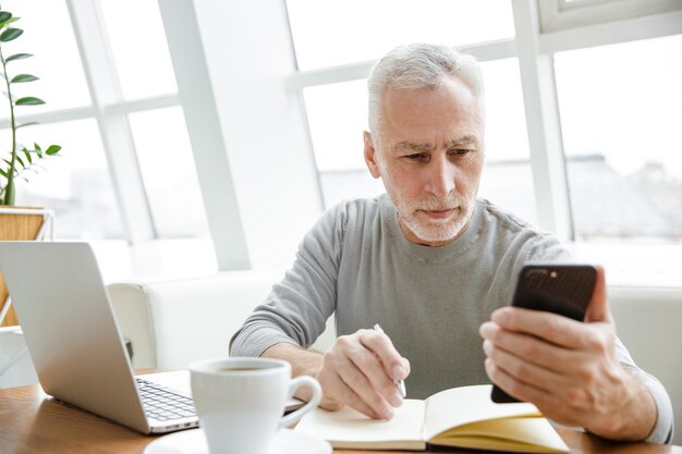 Ernster reifer Mann, der sich Notizen macht, während er mit Laptop und Handy im Café drinnen arbeitet?