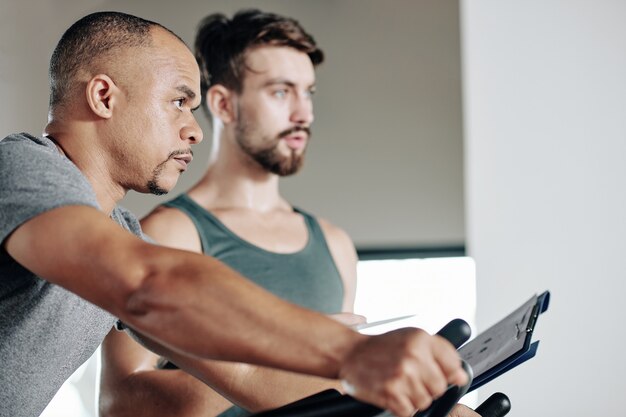 Ernster reifer Mann mit gemischter Rasse, der auf einem stationären Fahrrad im Fitnessstudio fährt, sein Trainer steht in der Nähe