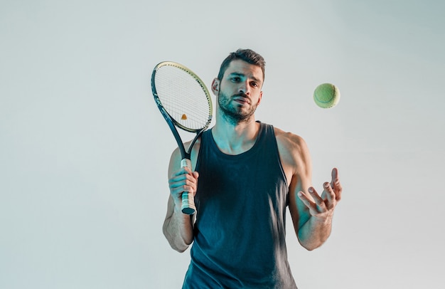 Ernster Tennisspieler hält Schläger und wirft Ball hoch. Vorderansicht des jungen bärtigen europäischen Sportlers, der die Kamera betrachtet. Auf grauem Hintergrund mit türkisfarbenem Licht isoliert. Studio-Shooting. Platz kopieren