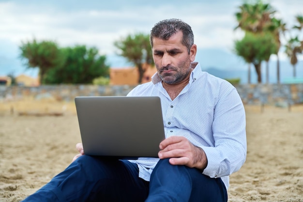 Ernster trauriger reifer Mann mit dem Laptop, der auf bewölktem Strand sitzt