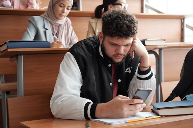 Ernster Typ, der Smartphone benutzt, während er unter Gruppenmitgliedern am Schreibtisch sitzt