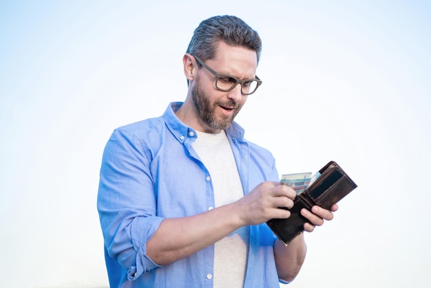 Ernster Typ mit Brille, der Bargeld aus dem Himmelshintergrund der Brieftasche nimmt