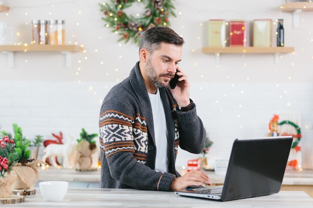 Ernster und selbstbewusster Mann, der zu Hause an einem Laptop arbeitet und während der Neujahrs- und Weihnachtsferien auf einem Mobiltelefon spricht