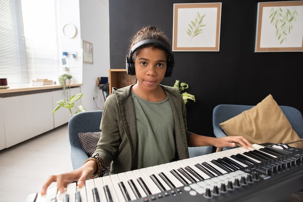 Ernstes afroamerikanisches Schulmädchen, das während der Wiederholung an der Klaviertastatur sitzt und dich ansieht