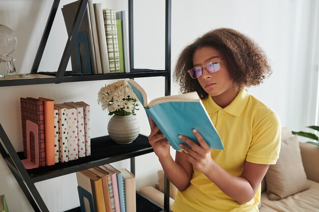 Ernstes gemischtes Teenager-Mädchen in Brille, das auf dem Sofa-Ellbogen sitzt und ein Buch in der Nähe des Regals im Wohnzimmer liest?
