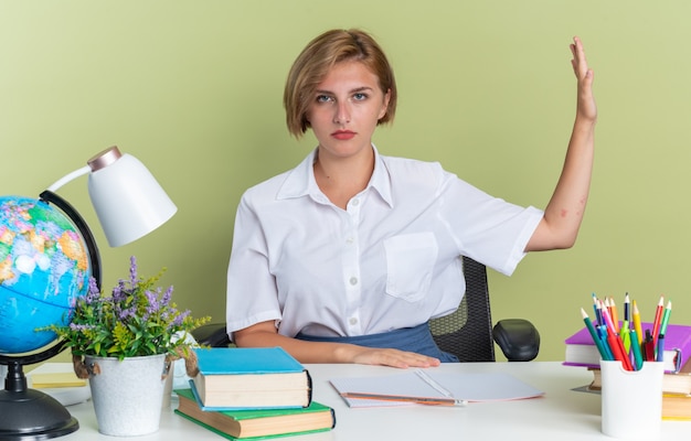 Ernstes junges blondes Studentenmädchen, das am Schreibtisch mit Schulwerkzeugen sitzt und in die Kamera schaut und die Stoppgeste zur Seite macht, isoliert auf olivgrüner Wand?