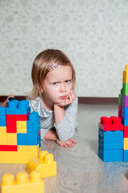 Ernstes Kindermädchen, das nahe hellen Plastikbausteinen liegt. Kleinkind spielt auf dem Boden. Spielzeug entwickeln. Frühes Lernen.