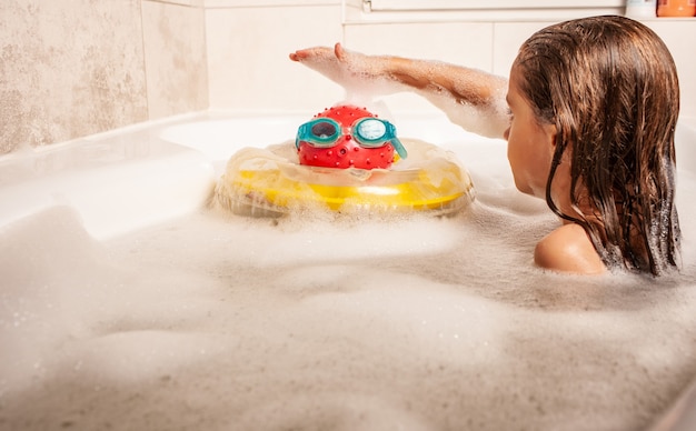 Ernstes kleines süßes Mädchen badet in einem Schaumbad und spielt mit Spielzeugen, die sich zum Meer vorstellen