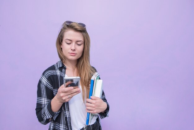 Ernstes Mädchen, das eine Freizeitkleidung trägt, benutzt ein Smartphone und hält die Notizbücher und Bücher auf dem lila Hintergrund.