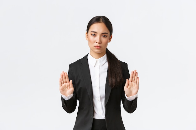 Ernsthaft aussehende unzufriedene asiatische Managerin, Geschäftsfrau im schwarzen Anzug, stoppt die Aktion, verbietet oder beschränkt etwas, hebt die Hände in einer verbietenden Geste, sagt nein oder genug, weißer Hintergrund