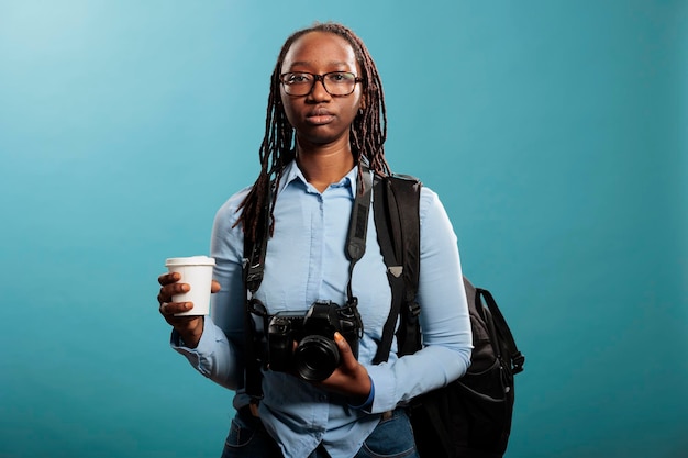 Ernsthaft aussehender professioneller Fotograf mit DSLR-Gerät und Tasse Kaffee, während er auf blauem Hintergrund in die Kamera schaut. Junger Fotografie-Enthusiast mit Fotoausrüstung, während er ein Getränk genießt.