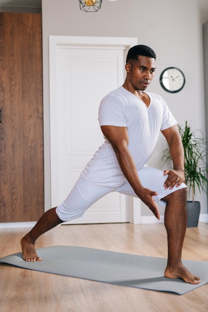 Ernsthaft fitter afroamerikanischer Mann, der Sport-Fitness-Übungen macht, die auf einer Yogamatte im hellen Wohnraum stehen und wegschauen. Konzept des Sporttrainings im Fitnessstudio zu Hause.