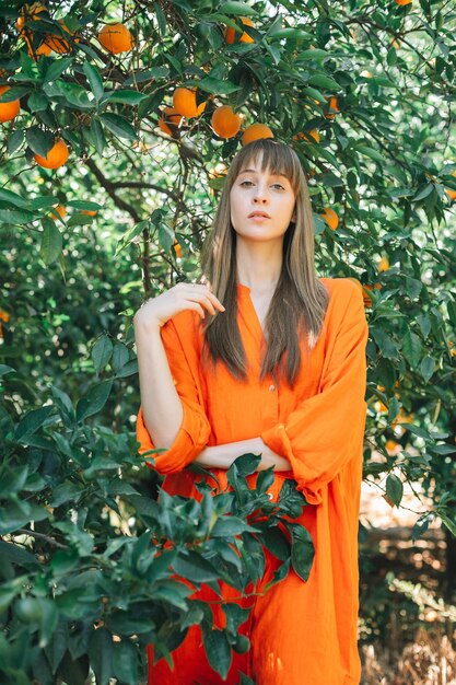 Ernsthaft junges Mädchen in orangefarbenem Kleid posiert vor der Kamera, indem es ihr Haar im orangefarbenen Garten hält