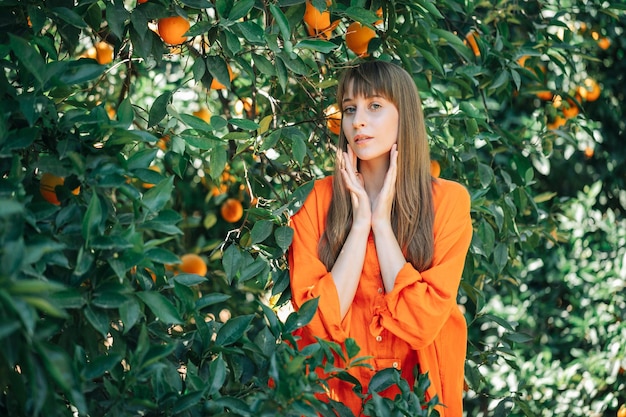 Ernsthaft junges Mädchen in orangefarbenem Kleid posiert vor der Kamera, indem es im Orangengarten die Hände in der Nähe der Wangen hält