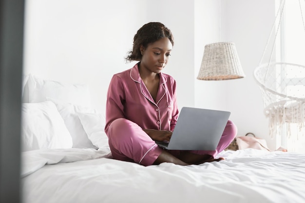 Ernsthafte afrikanische Dame mit Laptop, die im Schlafanzug im Schlafzimmer arbeitet