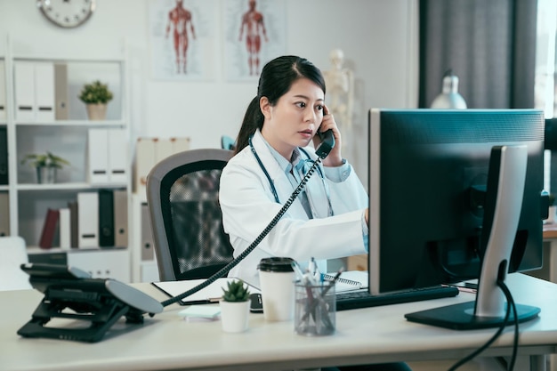 Ernsthafte asiatische Ärztin am Telefon im Büro. junge japanische ärztin im klinikkrankenhaus, die sich telefonisch mit der krankenschwester verbindet, um die patientensituation der operation auf dem computerbildschirm zu überprüfen