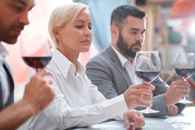 Ernsthafte blonde Frau, die Rotwein im Bokal betrachtet, während Farbe des Getränks zwischen ihren Kollegen bewertet