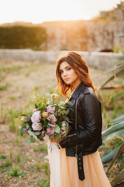 Ernsthafte Braut in einem Pastellkleid und einer schwarzen Lederjacke mit einem Blumenstrauß