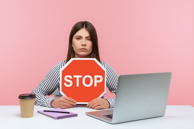 Ernsthafte Büroangestellte, die eine Stoppsymbol-Warnung mit rotem Verkehrsschild halten und wütend am Arbeitsplatz mit Laptop-Heimbüro sitzen Innenstudioaufnahme isoliert auf rosa Hintergrund