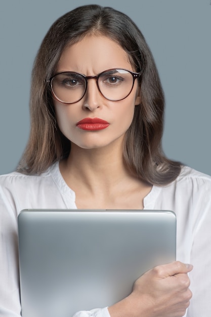 Ernsthafte Einstellung. Ernsthafte junge langhaarige Frau mit roten Lippen mit strengem Blick in der Brille, die Laptop fest an sie hält