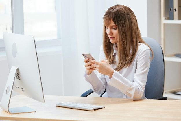 Ernsthafte, entzückende blonde Geschäftsfrau, Freiberuflerin, Blick auf das Telefon, das eine Nachricht in einem hellen, modernen Büro eingibt