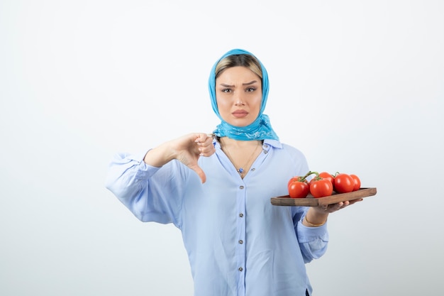 Ernsthafte Frau im blauen Schal, der Daumen nach unten zeigt und Holzbrett der roten Tomaten hält