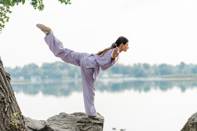 Ernsthafte Frau, Kungfu-Meisterin, trägt Kimono, trainiert, übt Wushu, steht auf einem Stein in der Nähe von Wasser, kopiert Raum, gesunder Lebensstil, Kampfkunstkonzept