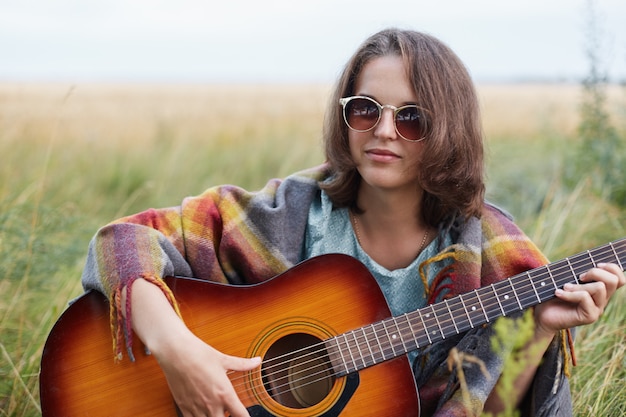 Ernsthafte Frau mit attraktivem Aussehen, die Sonnenbrille trägt, die auf Wiese mit akustischer Gitarre sitzt, die versucht, Lied zu spielen