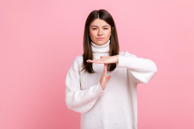 Ernsthafte Frau mit dunklem Haar, die eine Auszeitgeste zeigt, mit strengem Ausdruck, Frist, mit weißem Pullover im lässigen Stil. Studioaufnahme im Innenbereich isoliert auf rosa Hintergrund.