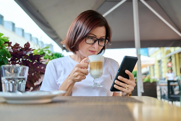 Ernsthafte Frau mittleren Alters, die in einem Café im Freien mit einer Tasse Kaffee sitzt und auf das Smartphone schaut