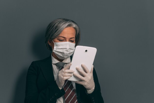 Foto ernsthafte geschäftsfrau im schwarzen anzug, in der schutzmaske und in den handschuhen, die mit digitalem gerät arbeiten. geschäft nach quarantänekonzept.